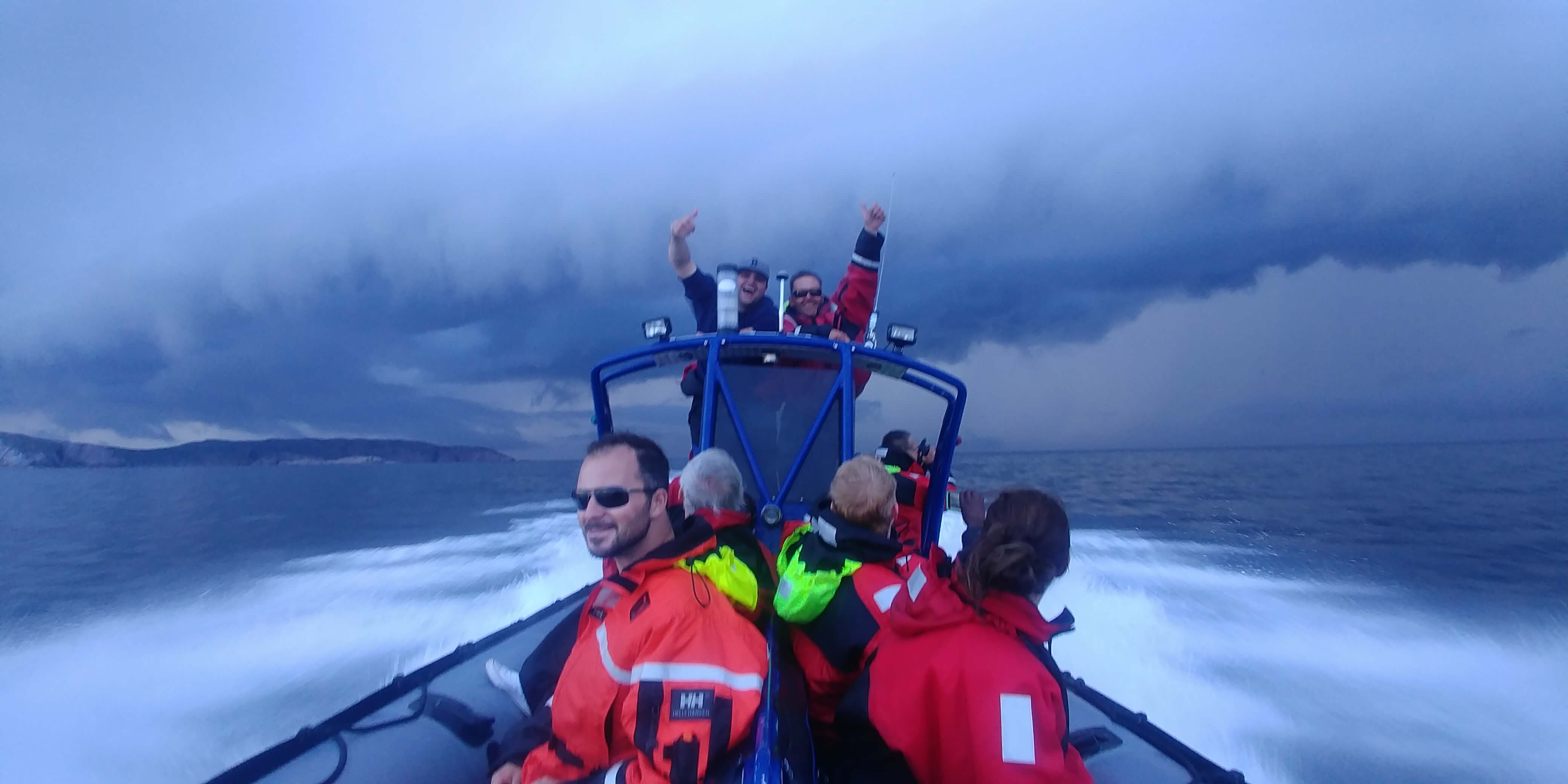 A group of people wearing flotation safety suits sit in a zodiac operated by Sea of Whales adventures