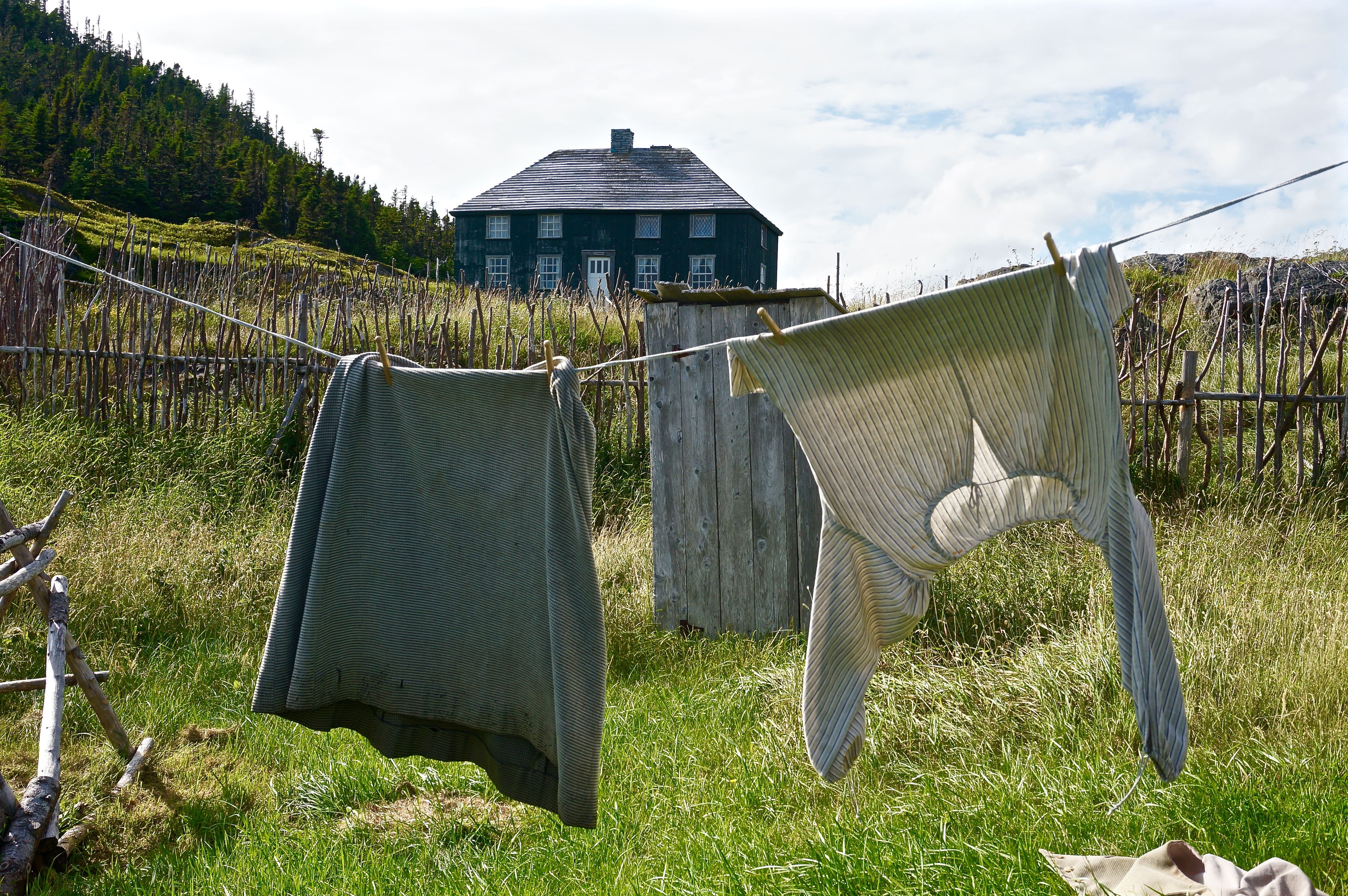A laundry line is hung with clothing from the 19th century with a square green house in the background