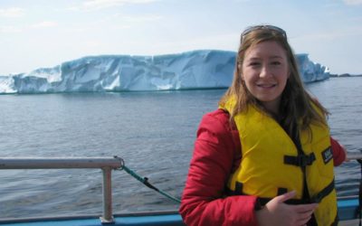 A passanger poses in front of an iceberg while on Rugged Beauty Boat Tour